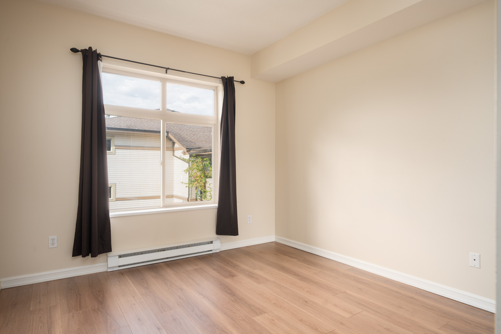 Empty room with beige walls, wood floor, black-curtained window. Sunlight reveals siding and roof outside in Surrey at 10088 148 St.