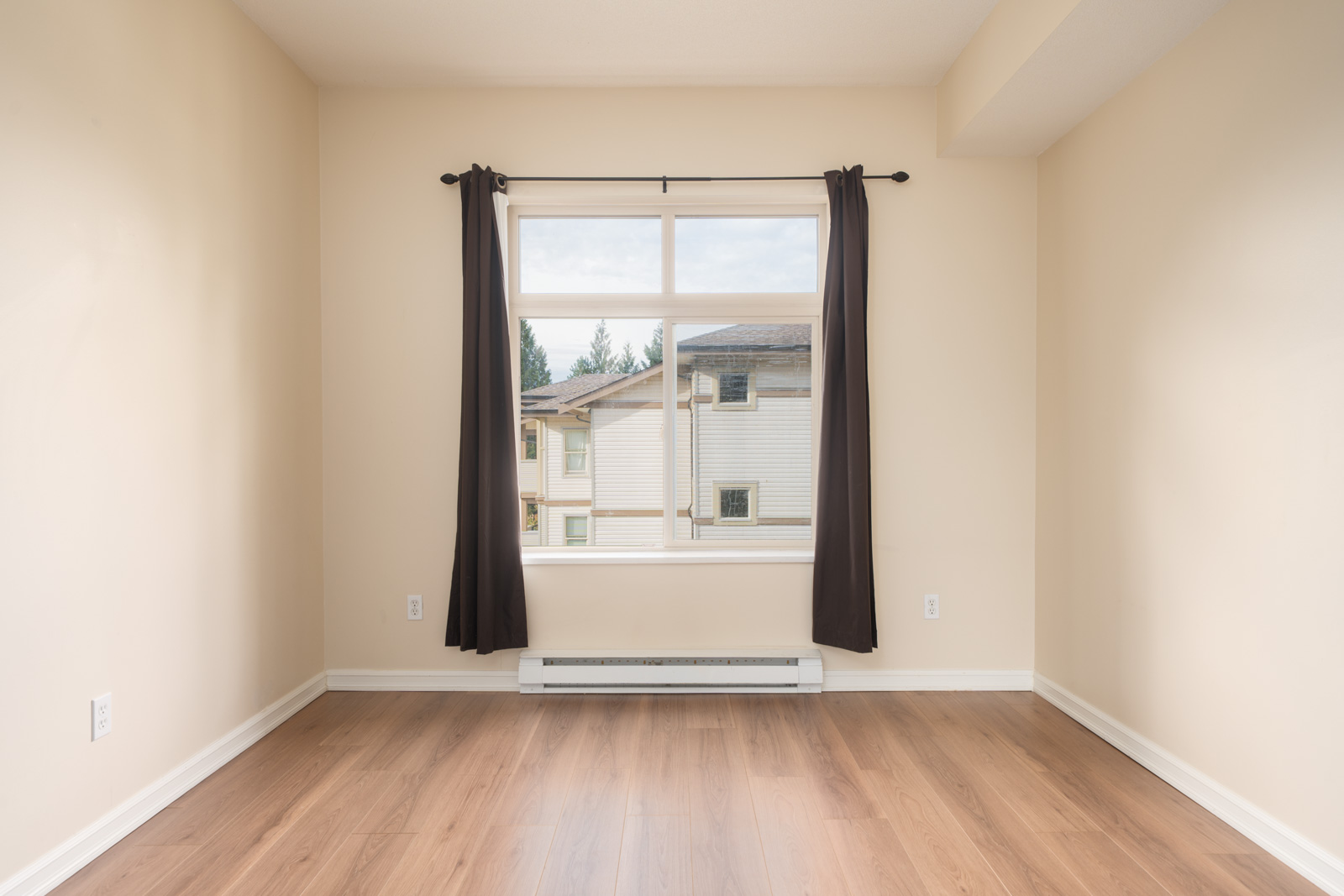 Empty room with beige walls, wooden floor, and a large window with dark curtains, facing buildings and trees in Surrey at 10088 148 St.