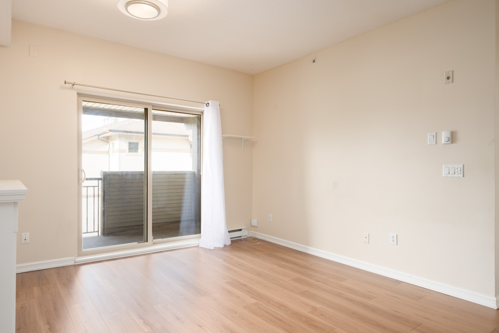 Empty room with light wood floors, beige walls, and a sliding glass door with white curtain to a small balcony at 10088 148 Street, Surrey.