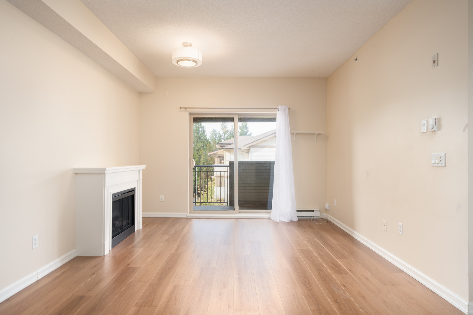 Empty living room in Surrey near 148 St, light wood floor, white fireplace, sliding glass door to small balcony, cream walls.