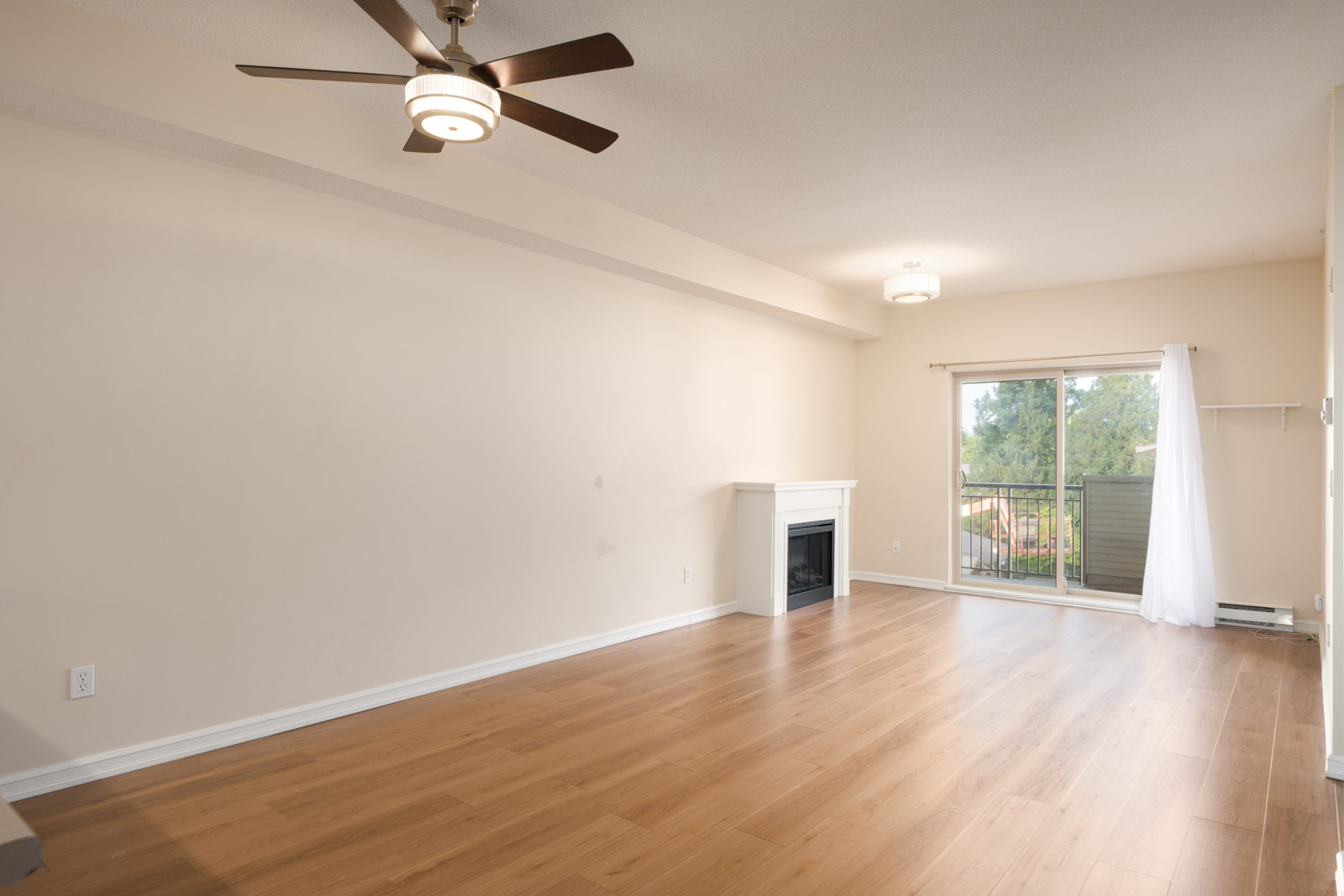 Unfurnished living room in Surrey with light wood floors, beige walls, ceiling fan, fireplace, and sliding door to balcony with tree view.
