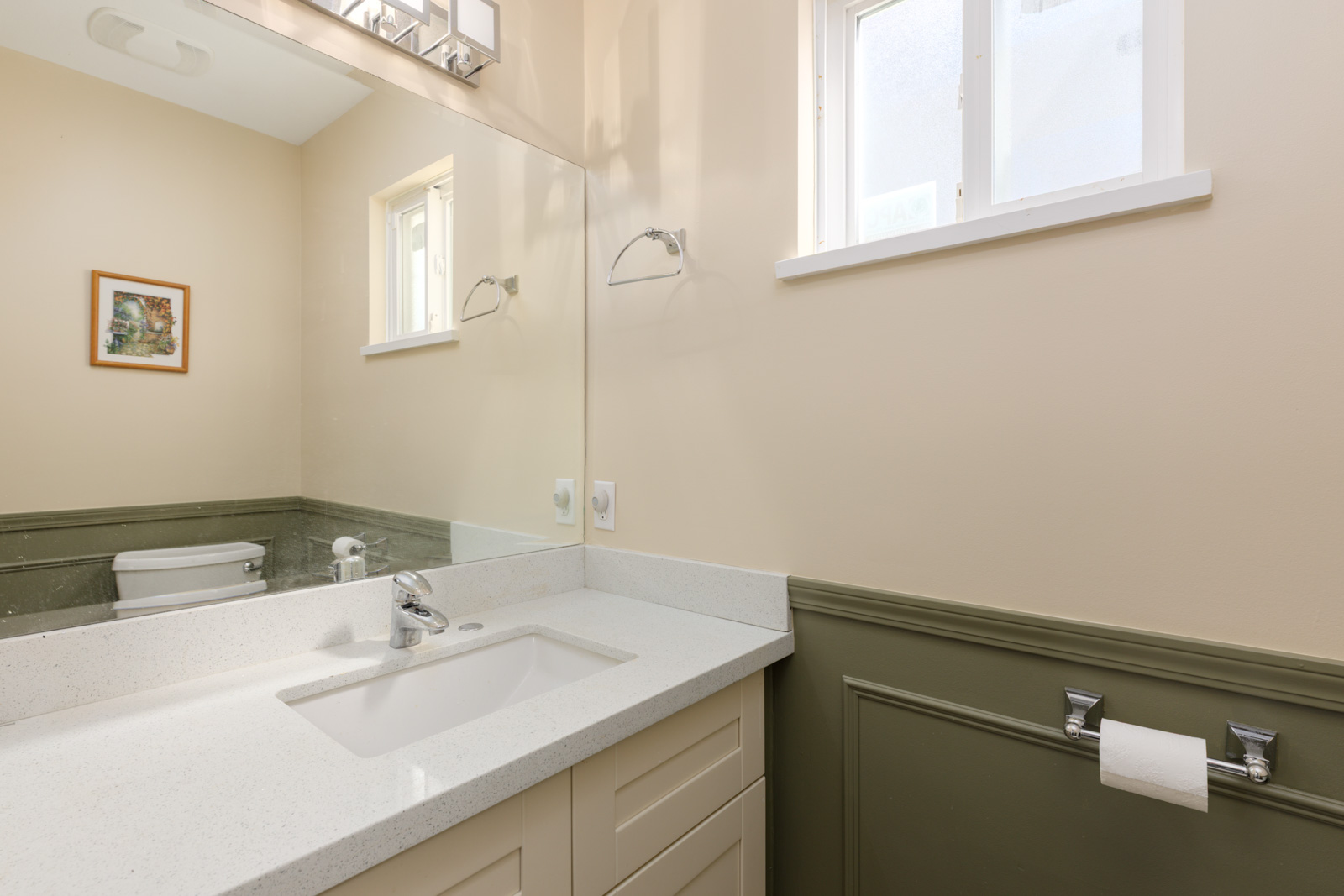 Bathroom with white sink, countertop, mirror, toilet, and toilet paper on green wainscoting; window above in Vancouver near Musqueamview St.