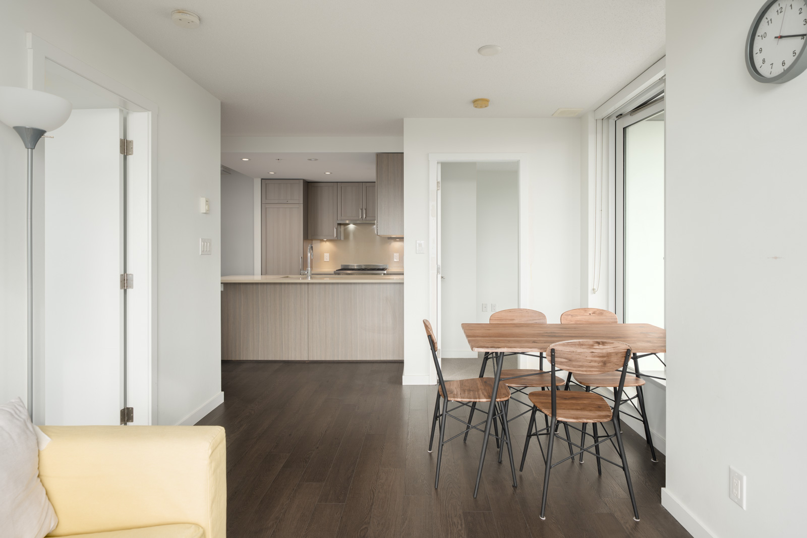 Vancouver apartment with white walls, dark wood floors, small dining set, open kitchen with light cabinets, and a wall clock.