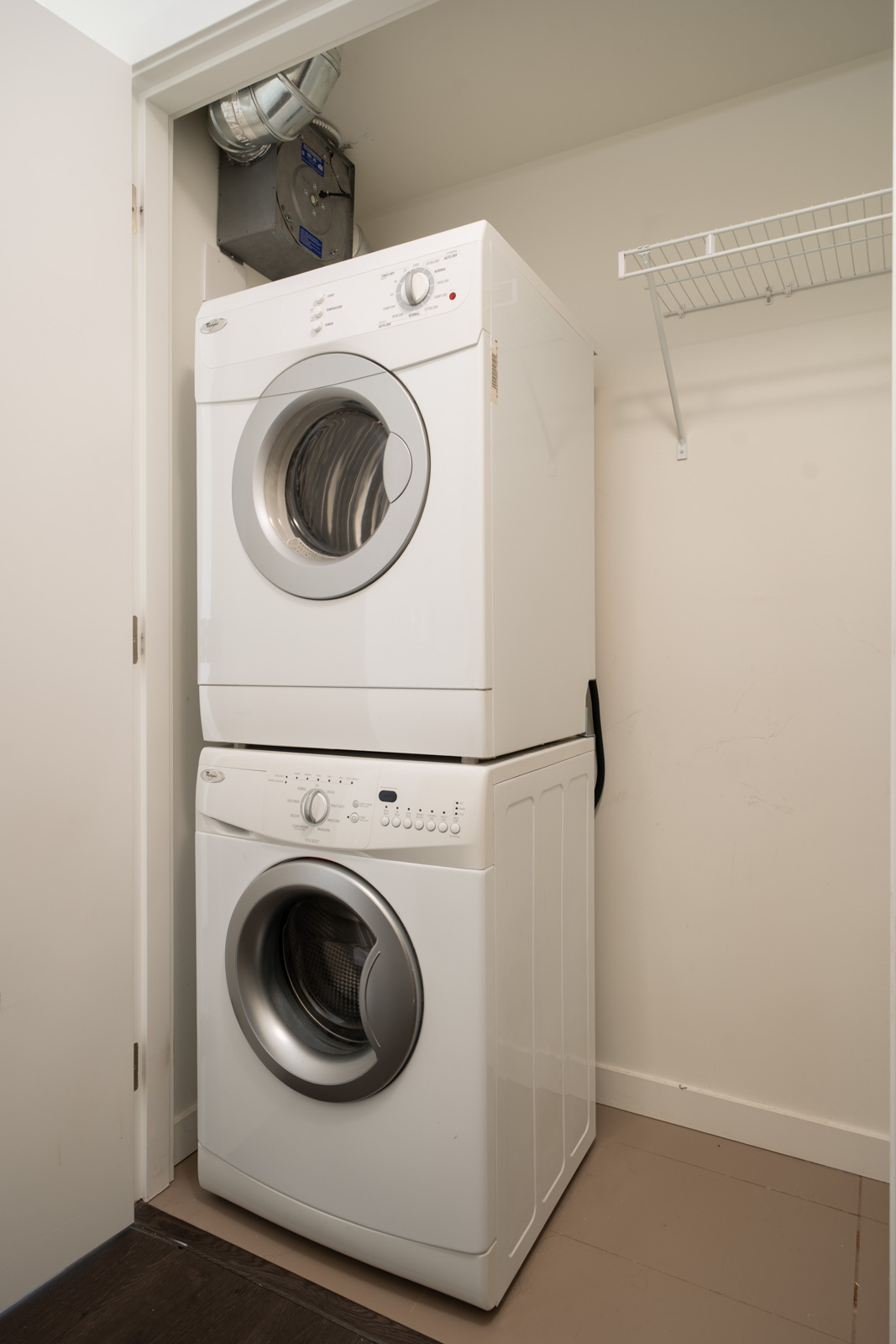 Stacked white washer and dryer in a compact laundry closet on Boundary Rd, Vancouver, with an empty wire shelf on the right.