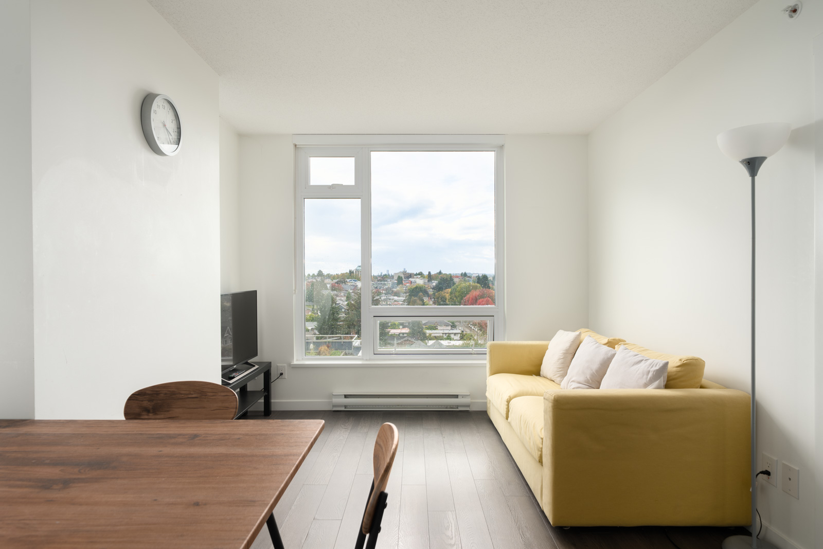 Minimalist living room with yellow sofa, wooden table and chairs, floor lamp, TV, and large window facing Boundary Rd, Vancouver.