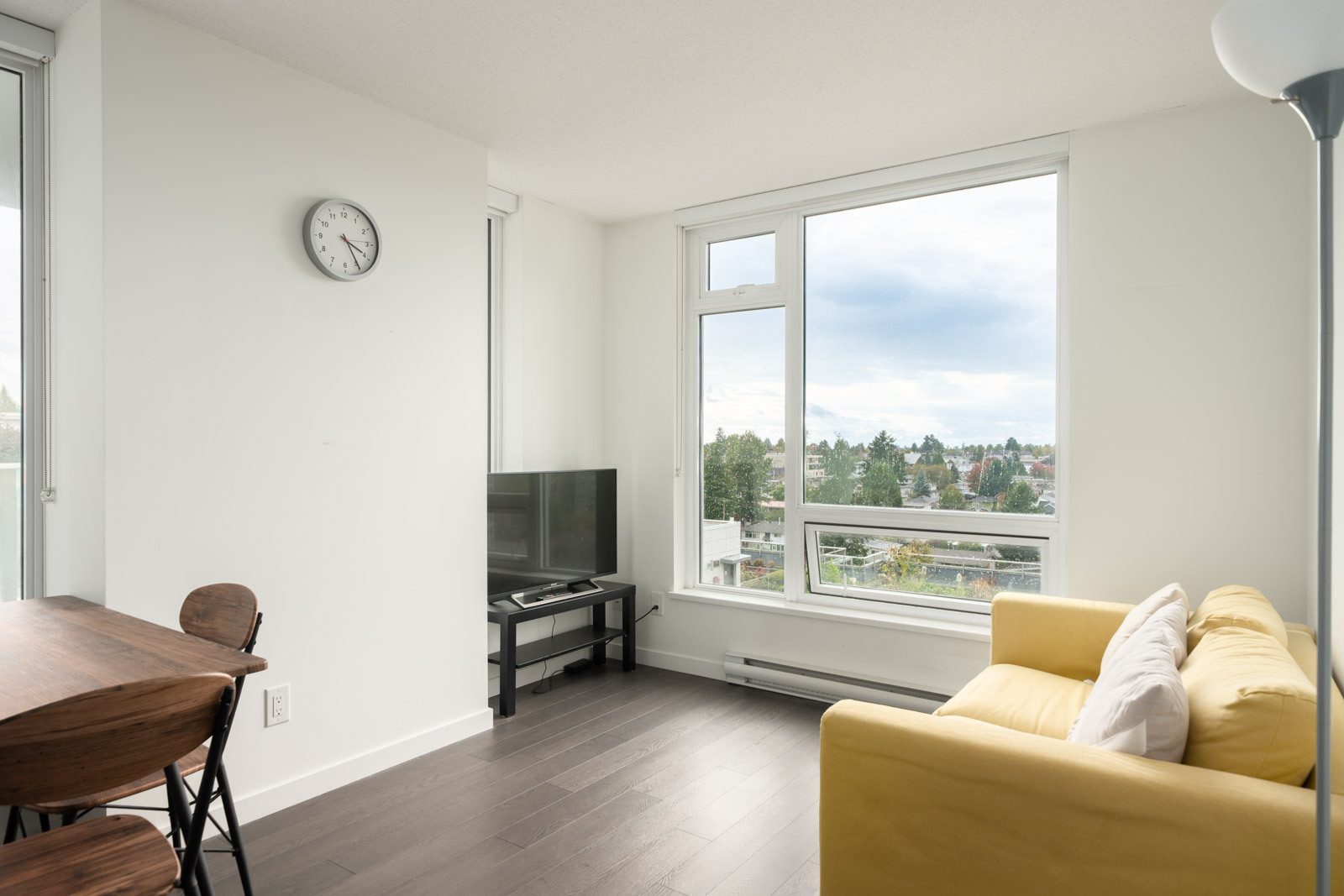 Bright living room in Vancouver with yellow sofa, TV stand, dining table, wall clock, and large windows showing trees and sky.