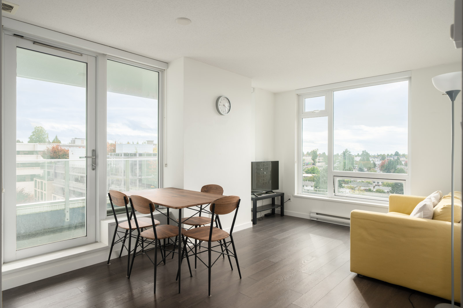 Vancouver apartment, 8th floor, large windows, yellow sofa, small dining table for four, TV on stand, clock, floor lamp, dark wood floor.