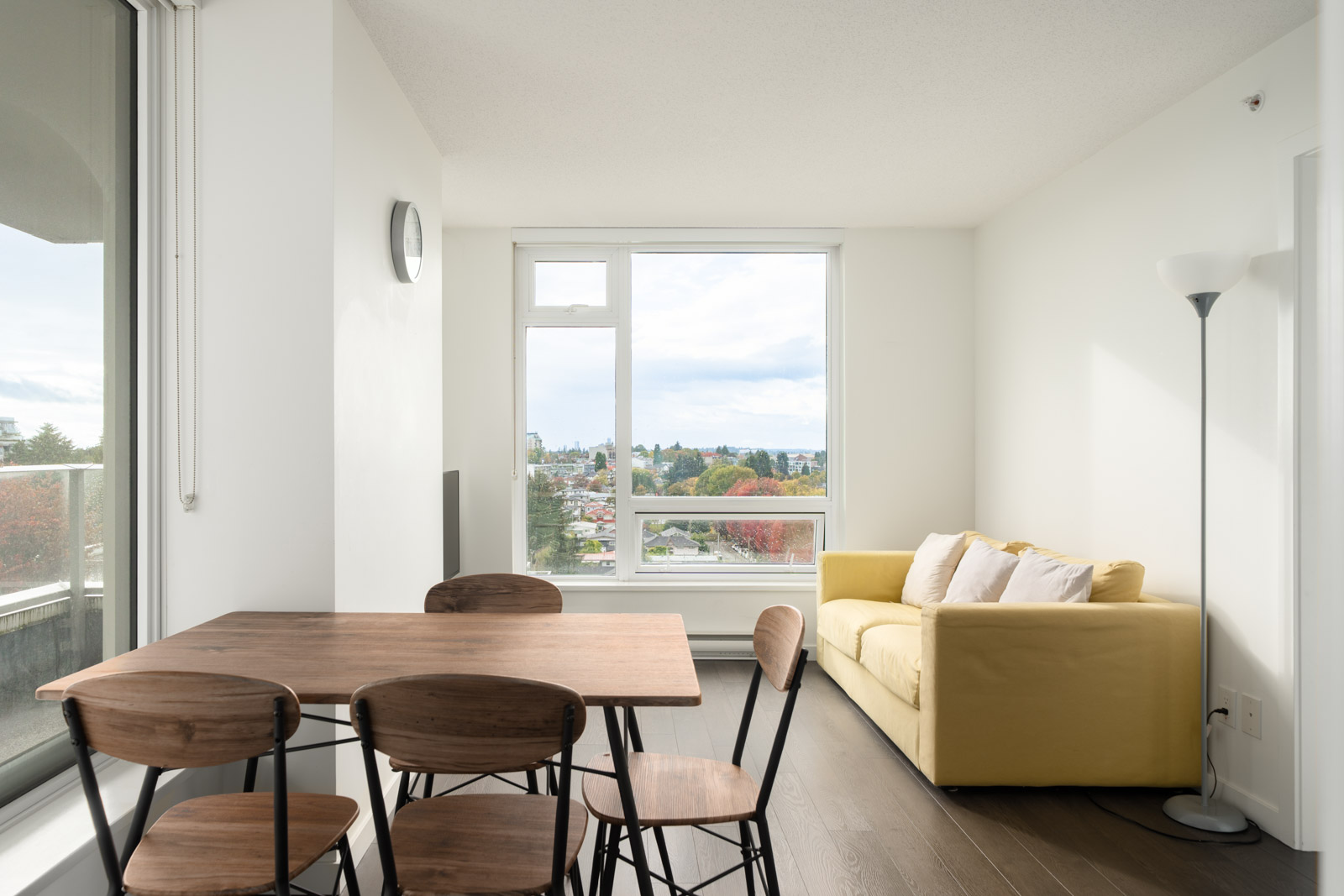 Modern apartment living space on 8th floor, Boundary Rd, Vancouver: wooden table, four chairs, yellow sofa, lamp, large city-view window.