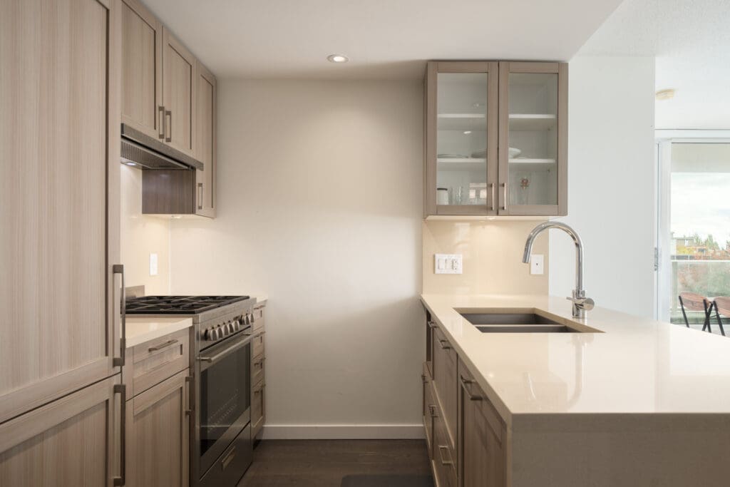 Modern kitchen with light wood cabinets, stainless appliances, beige counters, island sink, and sliding glass door in Vancouver.