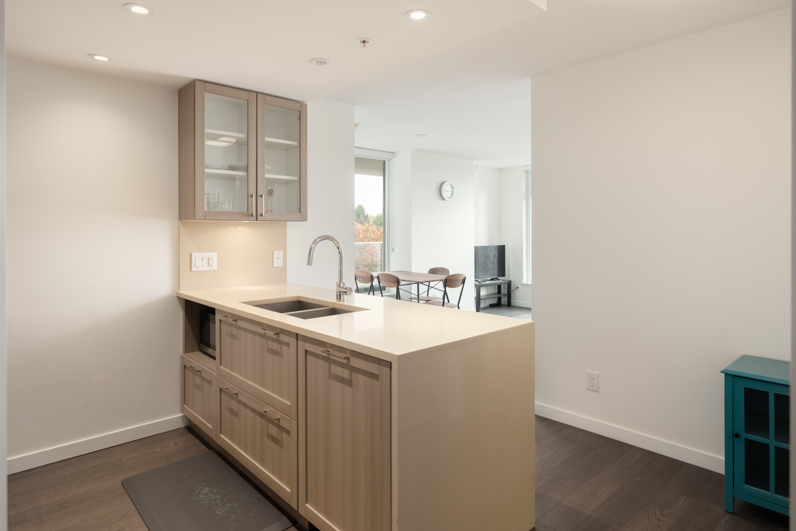Modern kitchen with beige cabinets and glass-front uppers, open to bright dining and living space with wall clock and TV, on Vancouver’s Boundary Rd, 8th floor.