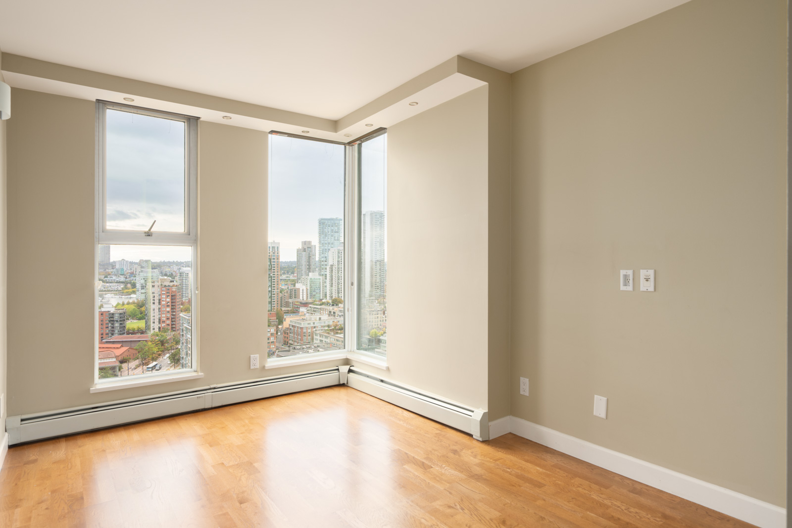 Bright corner room with light wood floors, beige walls, and large windows overlooking the Vancouver skyline at 1008 Cambie St.