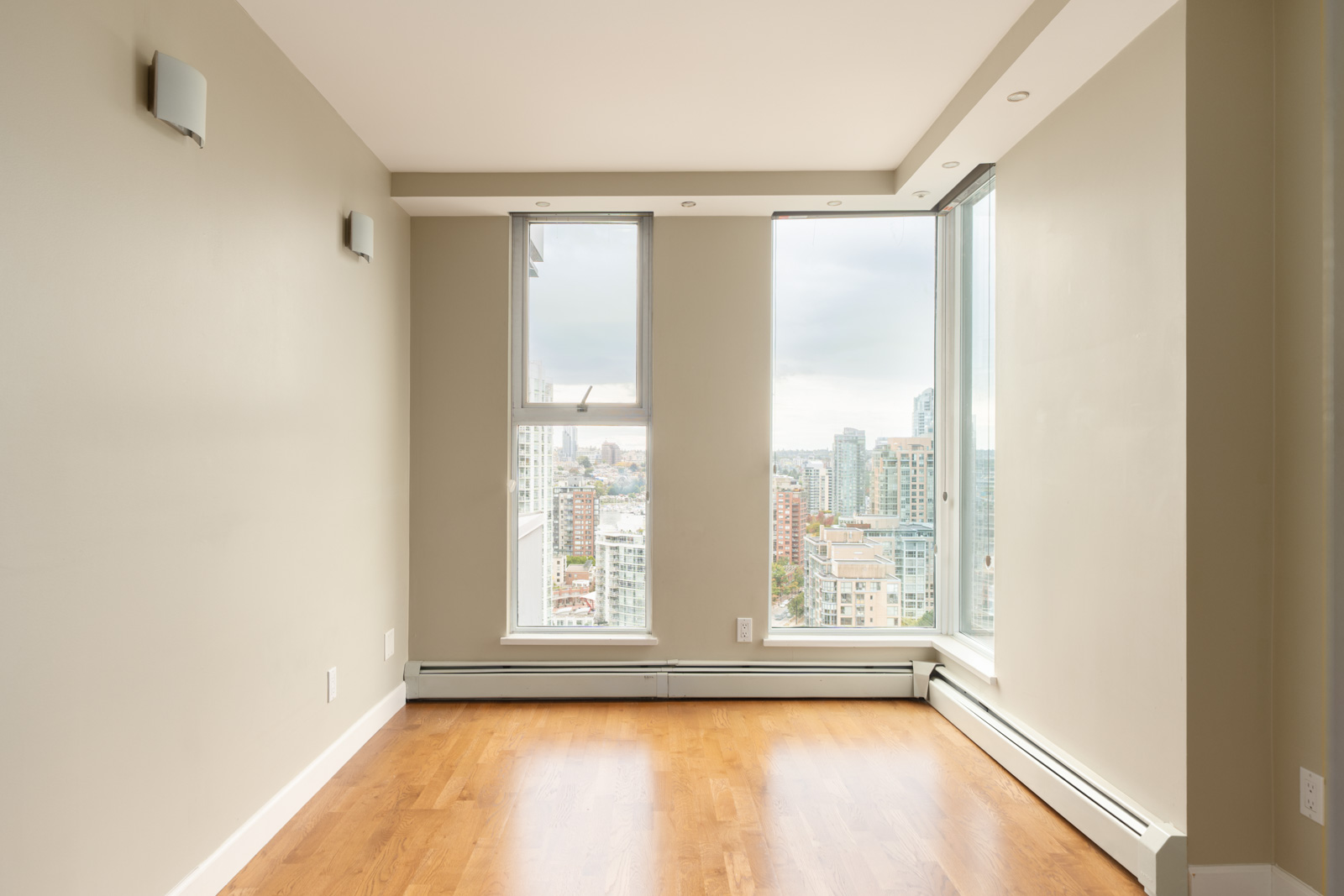 Empty room with light brown hardwood floors, beige walls, and large windows overlooking a cloudy cityscape in Vancouver.