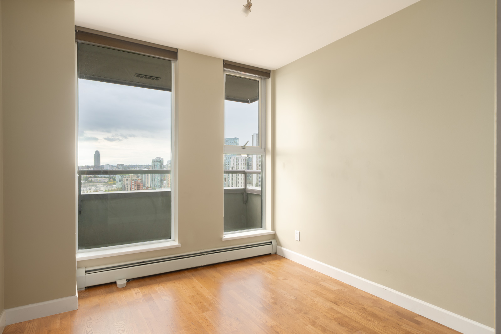 Empty room with light brown walls, wood flooring, two large windows showing a city view; baseboard heater under windows. Vancouver.