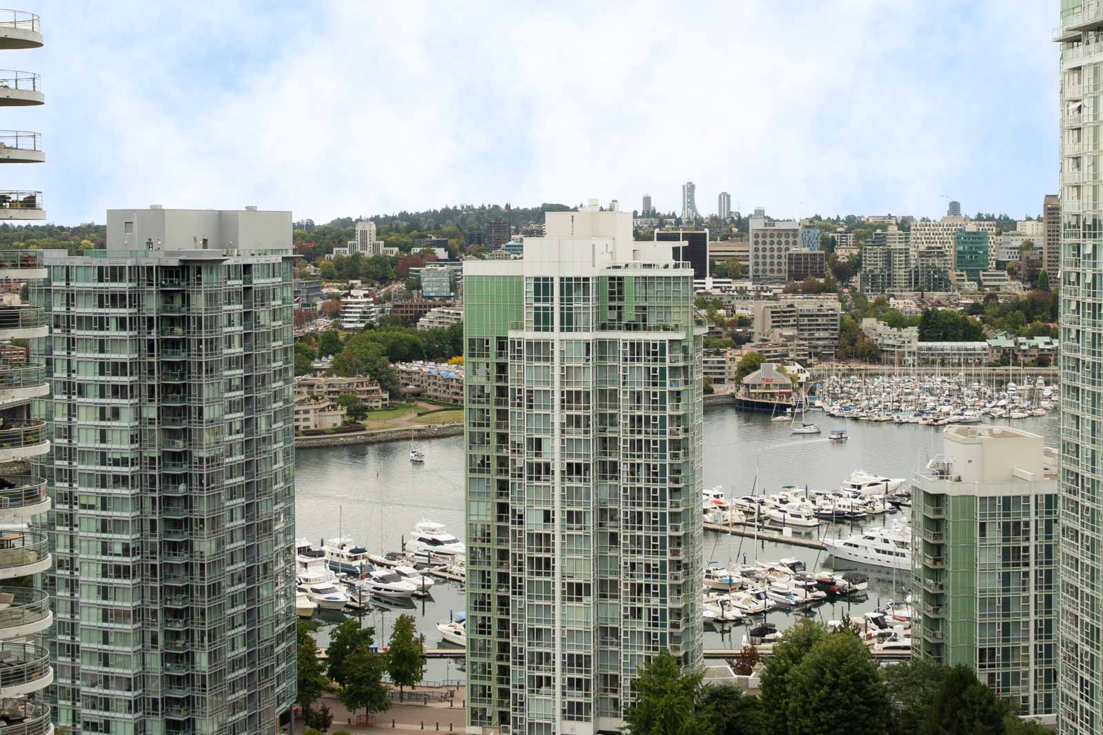 High-rise apartments overlook a boat-filled marina, with city buildings and trees in the background under partly cloudy Vancouver skies.