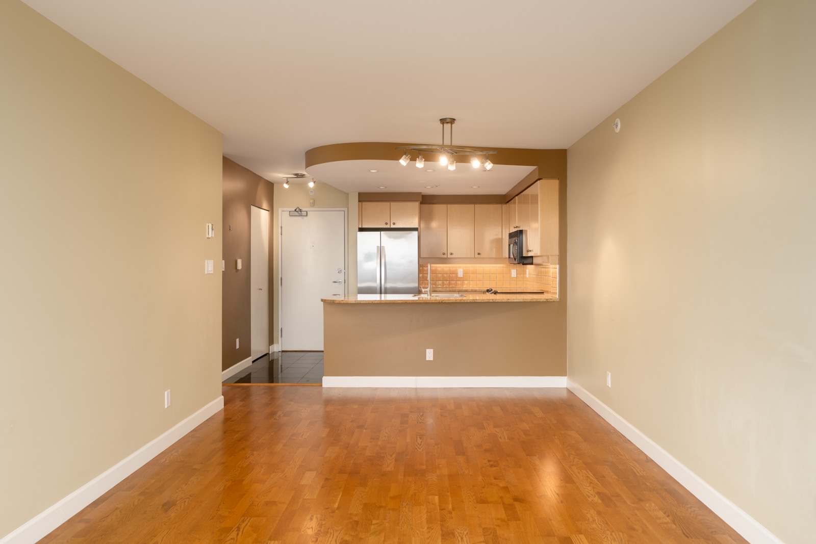 Unfurnished living area with hardwood floors and open kitchen, light wood cabinets, breakfast bar; white door left. Vancouver, 29th floor.
