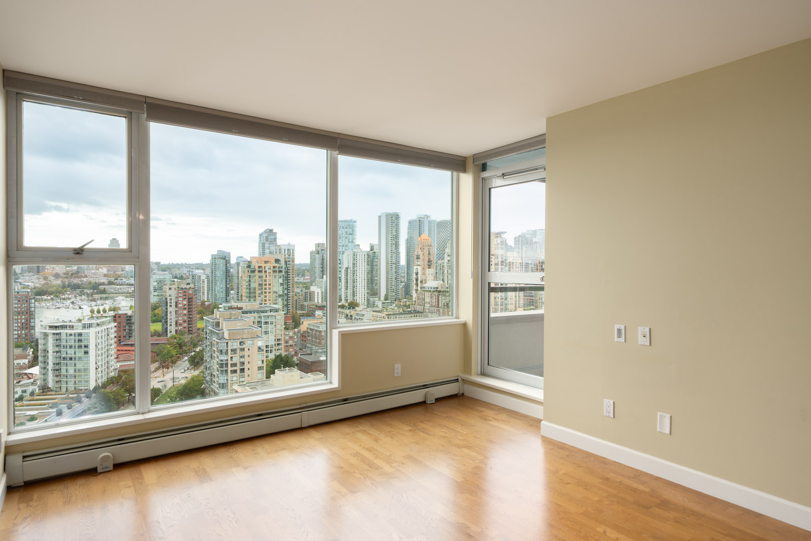 Empty room with hardwood floor, beige walls, large windows overlooking Vancouver skyline and a balcony through glass door at 1008 Cambie St.