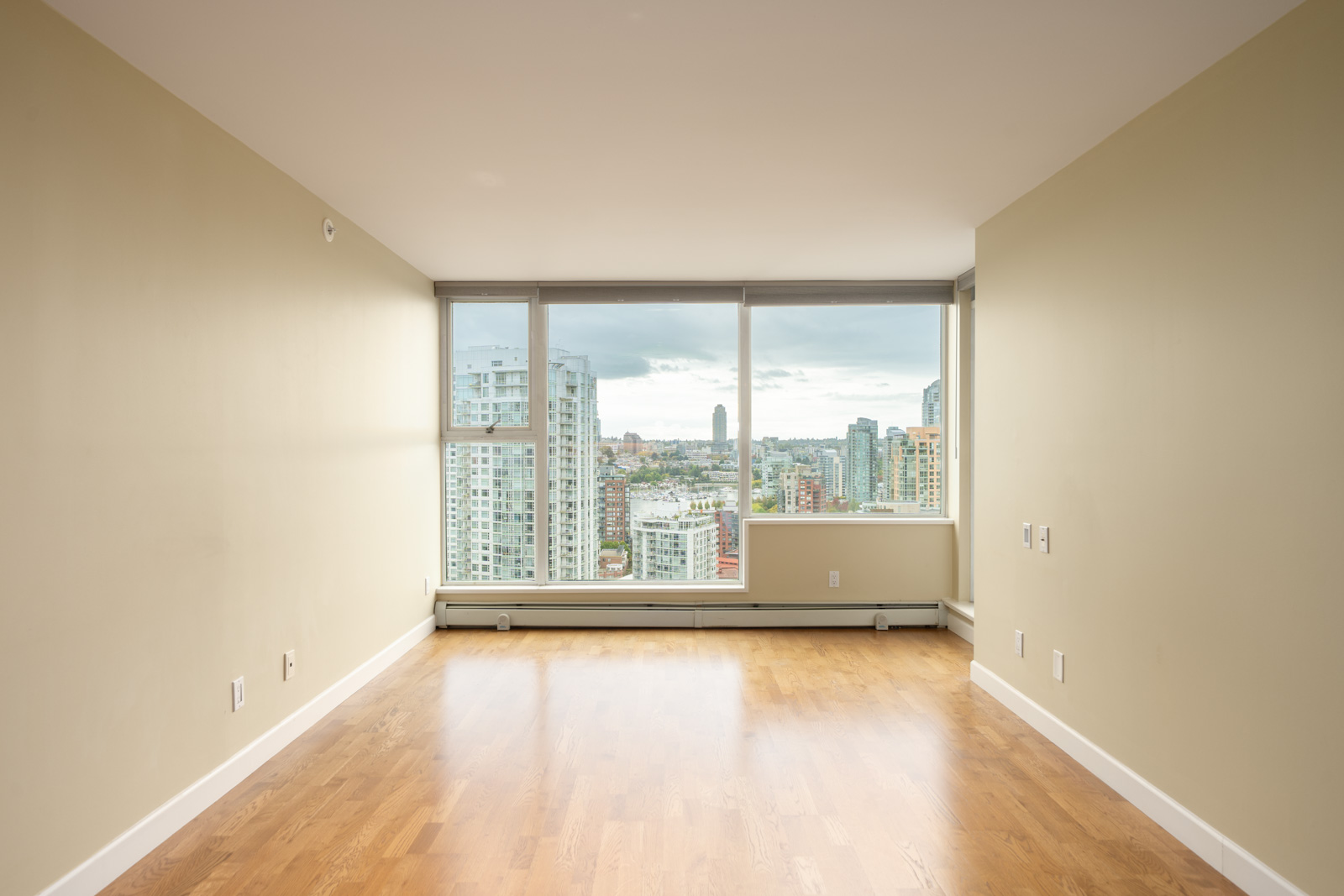 Empty room with beige walls, wood floors, and large windows at 1008 Cambie St, overlooking Vancouver’s skyline on a cloudy day.