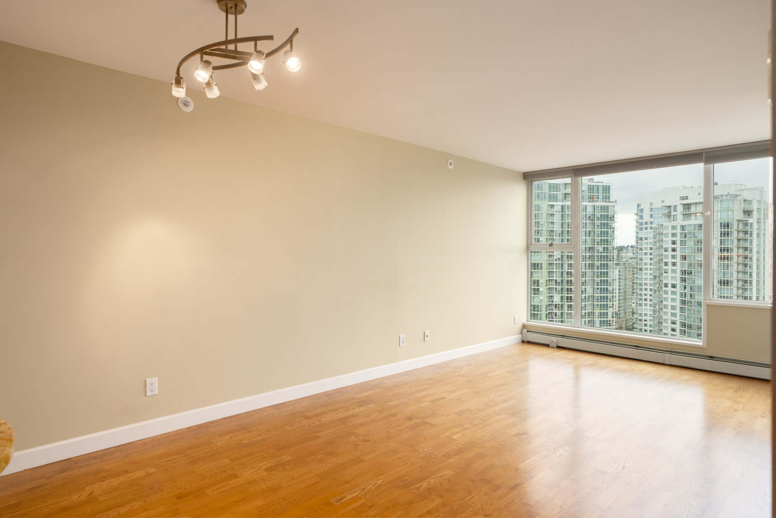 Empty living room at 1008 Cambie St, Vancouver, with light wood floors, beige walls, modern ceiling light, and large city-facing windows.