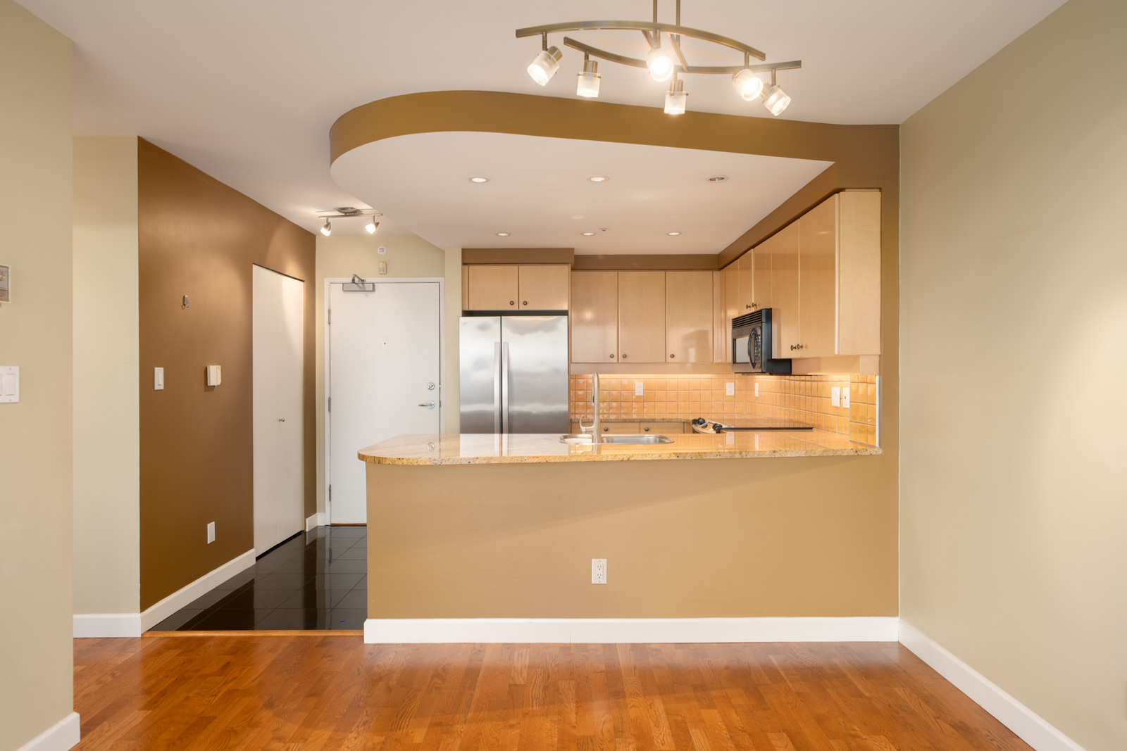 Modern kitchen with light wood cabinets, stainless appliances, granite counters, track lighting, and hardwood floors; entryway left.