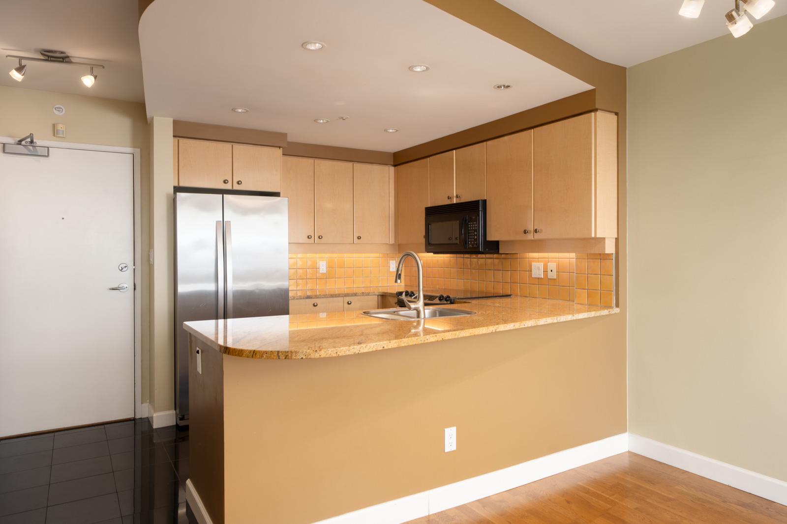 Modern kitchen on Vancouver’s Cambie St, 29th floor, with light wood cabinets, stainless fridge, yellow backsplash, granite counters.