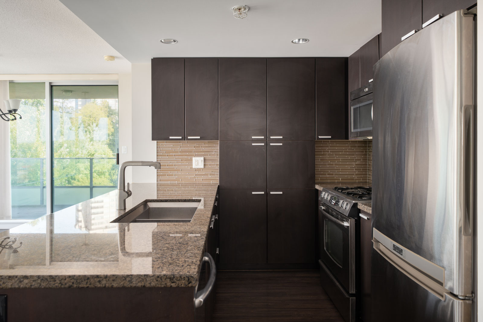 Modern kitchen with dark cabinets, stainless appliances, granite counters, tile backsplash, and large window on 7th floor in Vancouver.