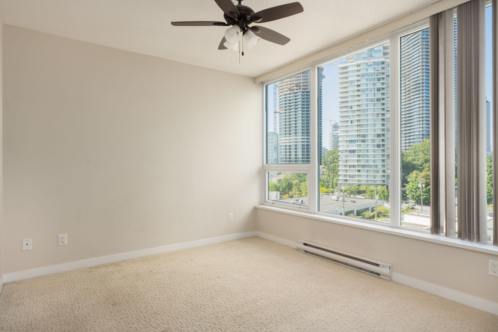 Empty room with beige carpet, cream walls, ceiling fan, large windows, and view of city buildings from 7th floor in Vancouver.