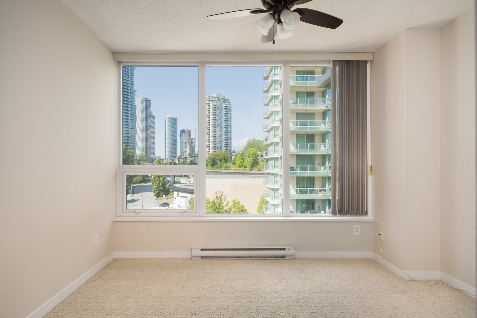 Empty room with beige carpet, large window, ceiling fan, and a city view of tall buildings on Douglas Rd in Vancouver, 7th floor.