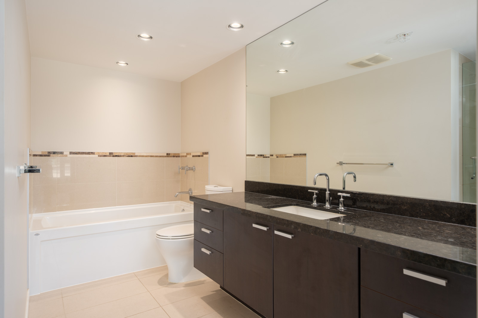 Modern Vancouver bathroom with bathtub, toilet, double-sink dark vanity, large mirror, beige tile floor, and recessed lighting.