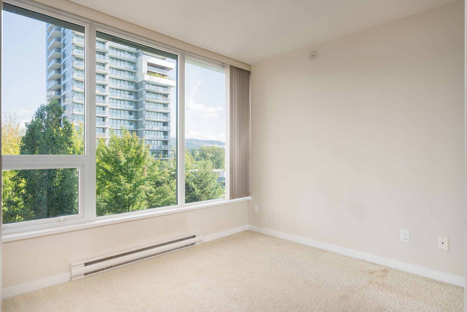 Empty room with beige carpet and white walls, large windows overlooking trees and a high-rise on the 7th floor in Vancouver.