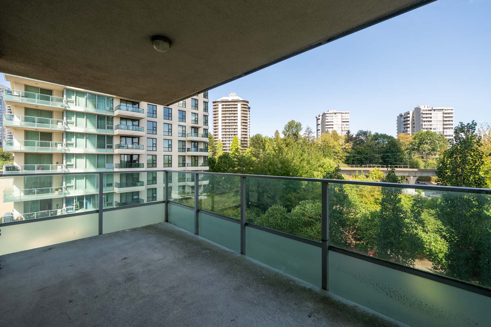 Covered balcony with glass railing on 7th floor in Vancouver, overlooking trees and mid-rise buildings along Douglas Rd on a clear day.