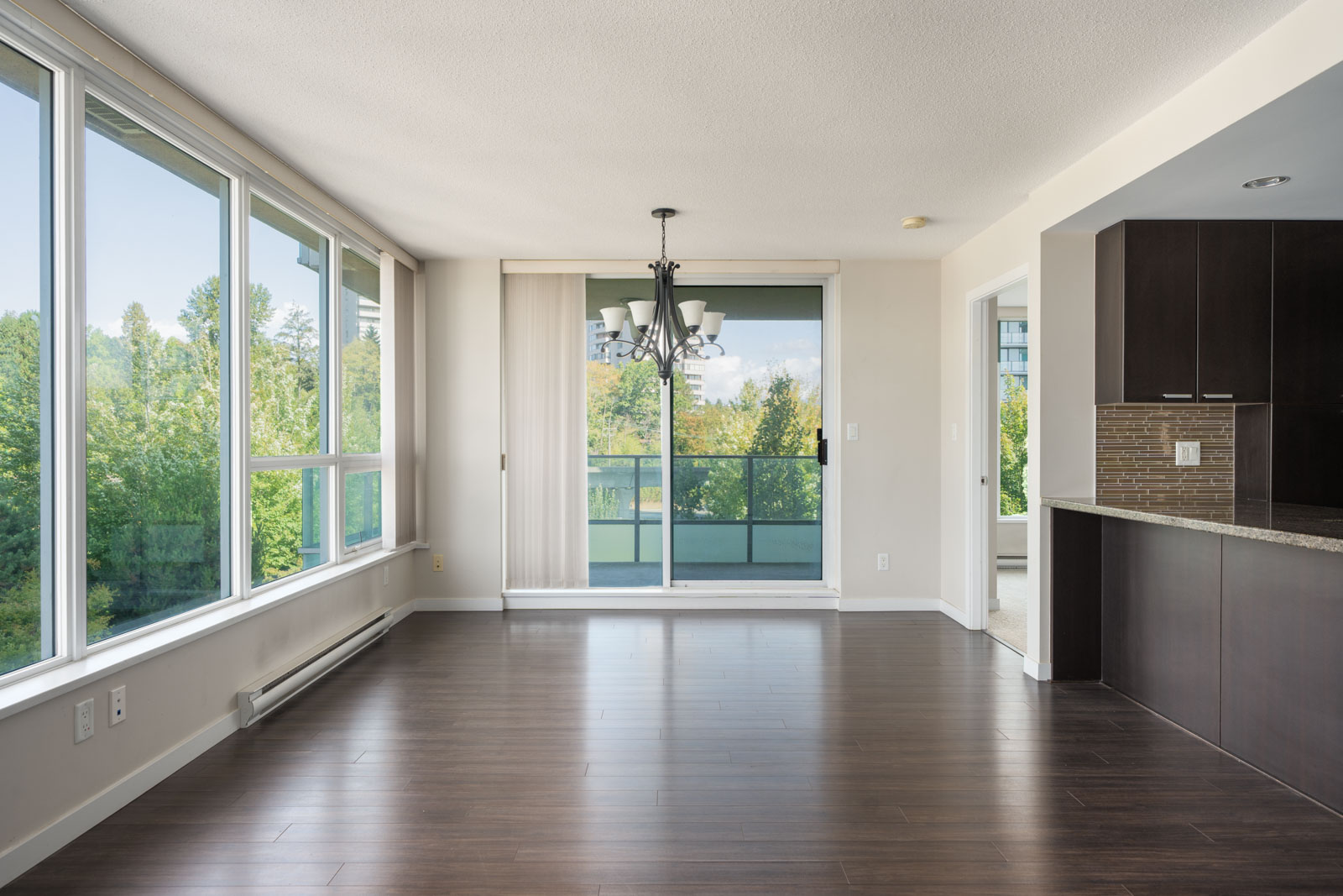 Empty modern apartment with large windows, dark wood floors, chandelier, and balcony doors overlooking greenery at 2232 Douglas Rd, Vancouver.