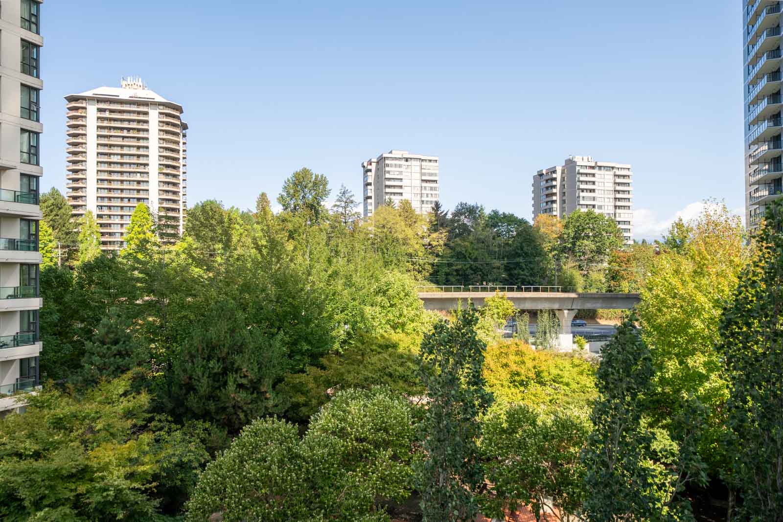 Four tall apartment buildings above dense green trees near Douglas Rd, Vancouver, with a concrete overpass and clear blue sky.