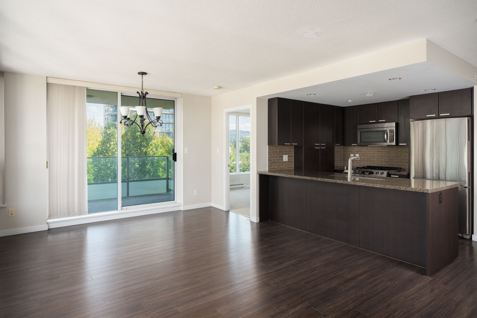 Empty modern apartment on 7th floor in Vancouver with dark wood floors, open kitchen, balcony, and view of green trees at 2232 Douglas Rd.