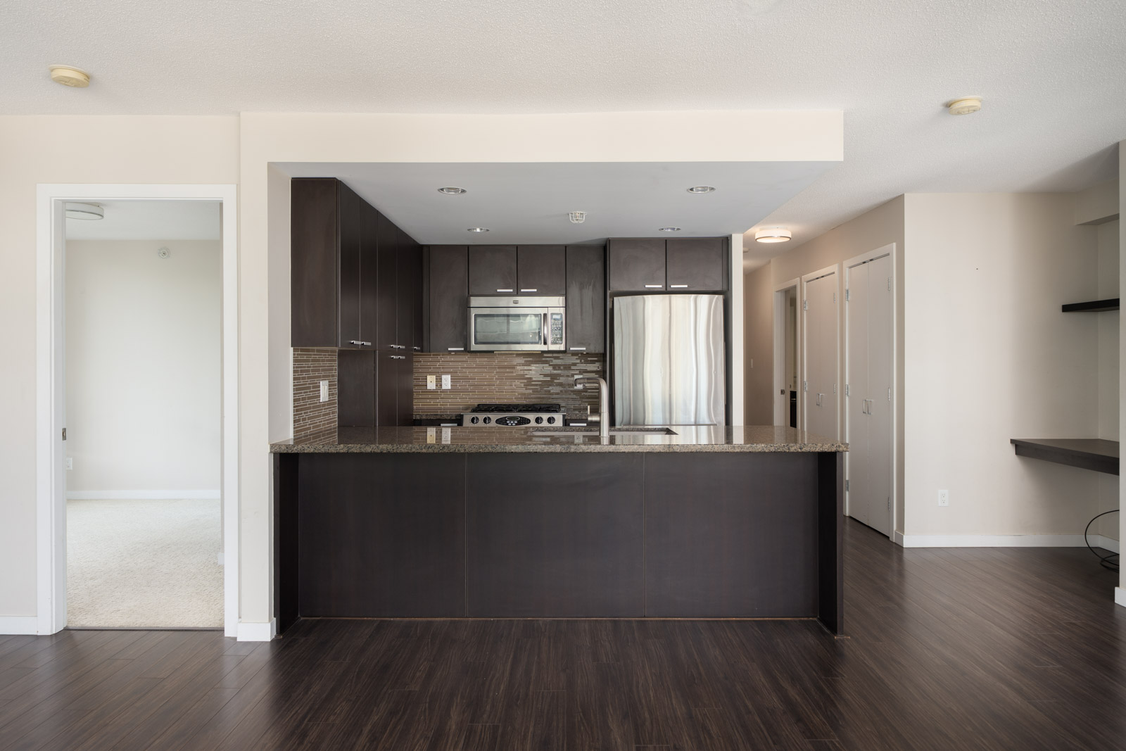 Modern kitchen with dark cabinets, stainless appliances, granite counters, wood floor; doors to carpeted room, Vancouver 7th floor.