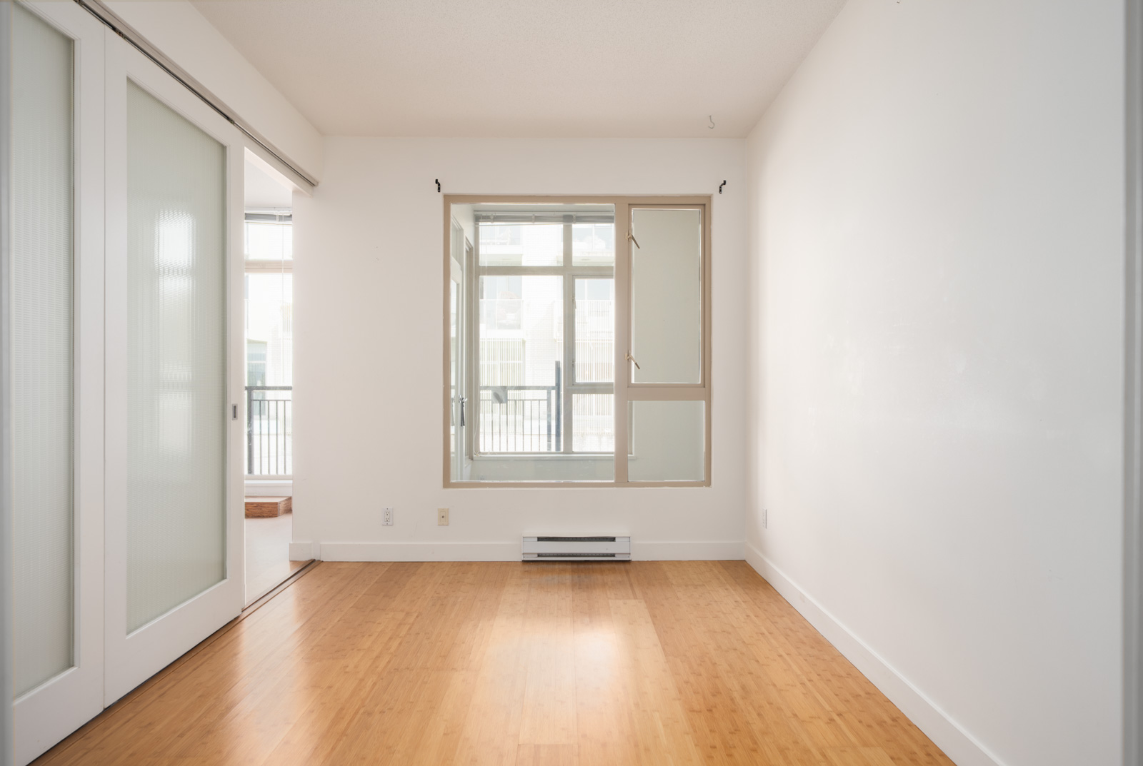 Empty room with light wood floors, white walls, large window with beige frame, and sliding doors on left, 4th floor, Vancouver.