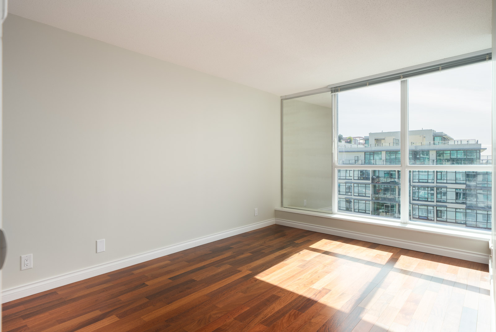 Empty room with light brown hardwood floors, off-white walls, and large windows overlooking North Vancouver cityscape.