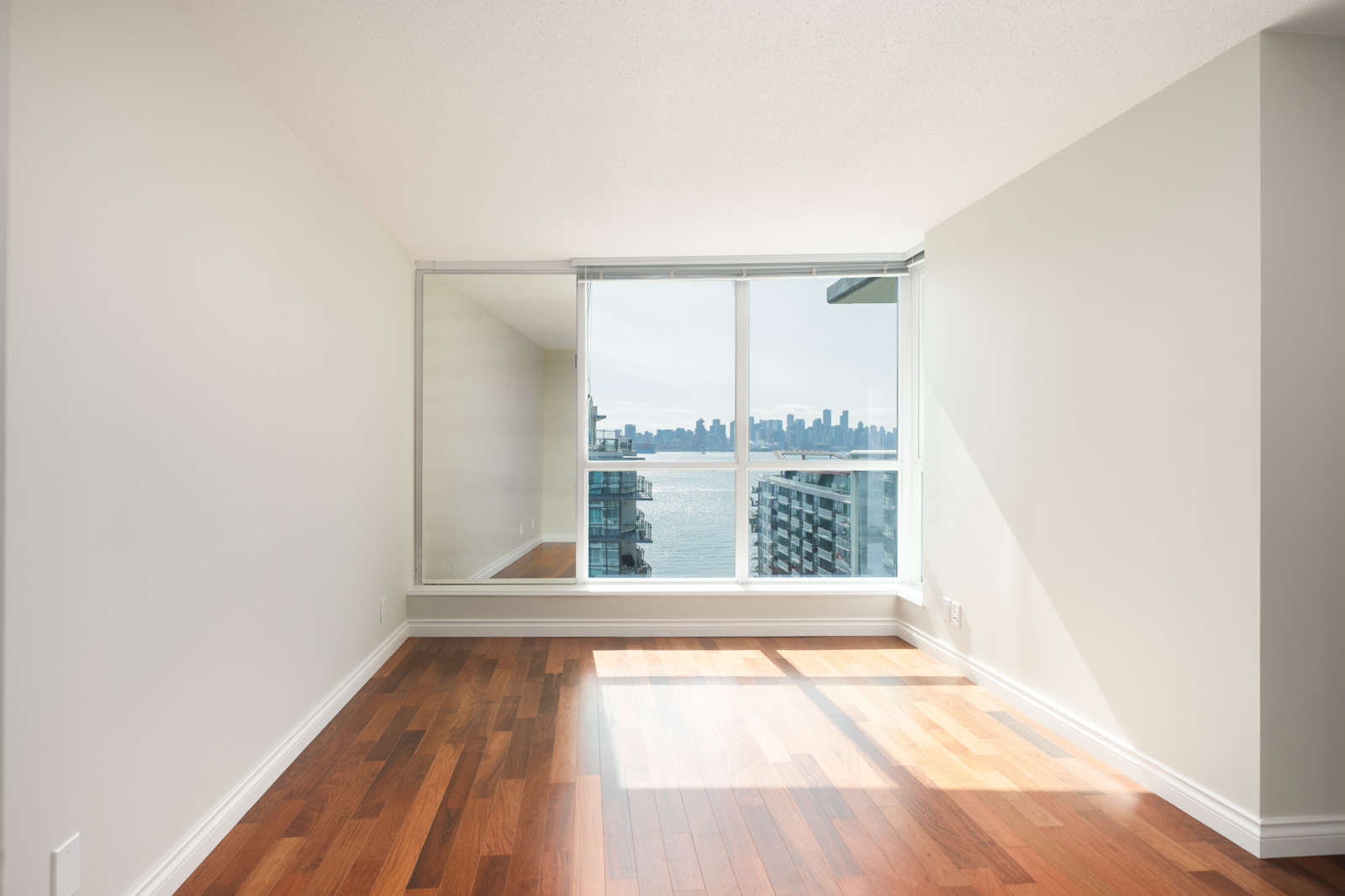 Bright, empty room on 12th floor at 188 E Esplanade, North Vancouver; hardwood floor, large window with water and city views, mirrored wall.