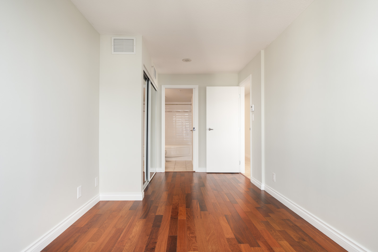 Empty room with white walls, wood floor, mirrored closet, and open door to bathroom with tub in North Vancouver high-rise.