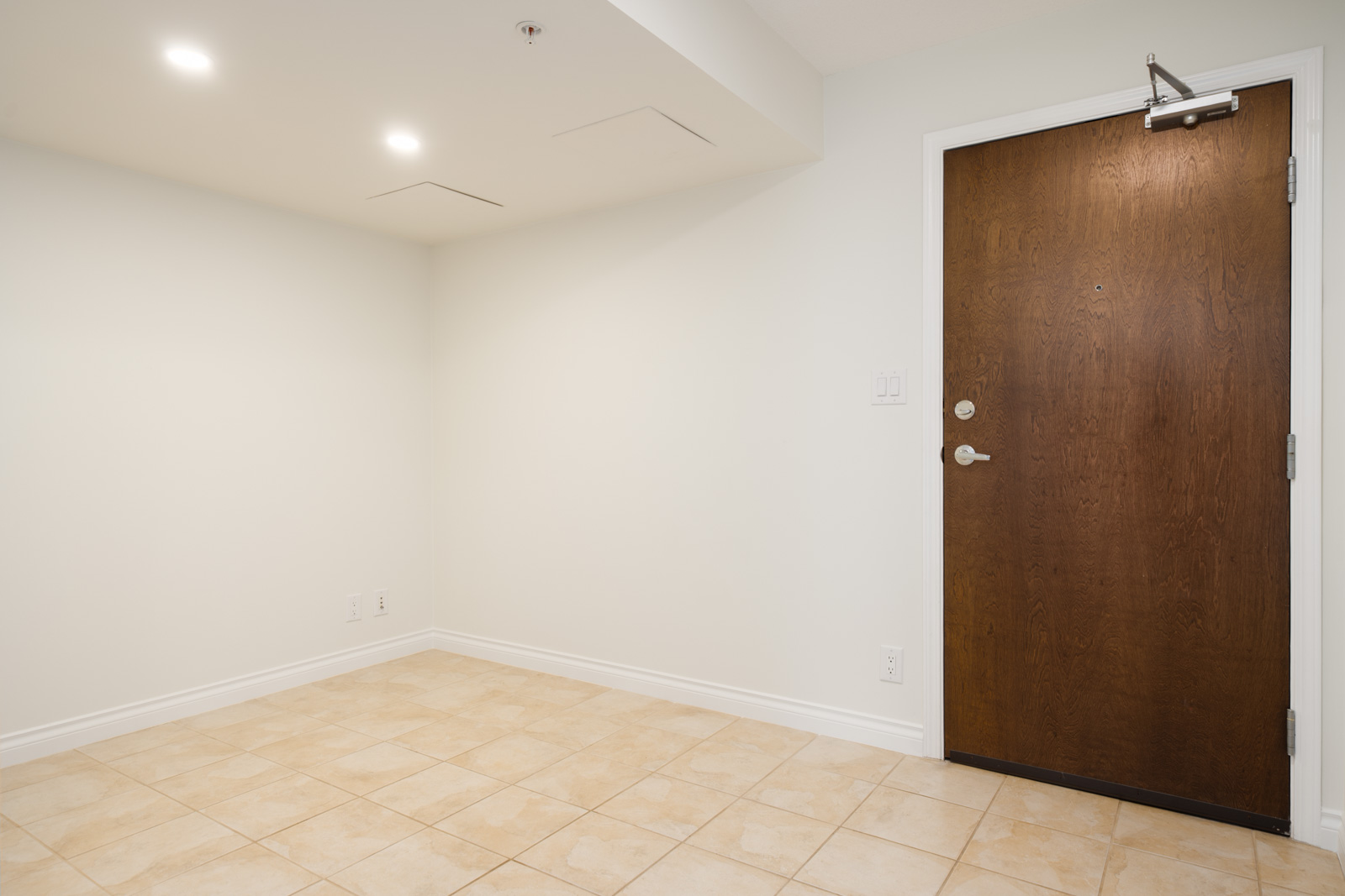Small empty room on the 12th floor at 188 E Esplanade, North Vancouver; beige tile floor, white walls, brown door, ceiling lights on.