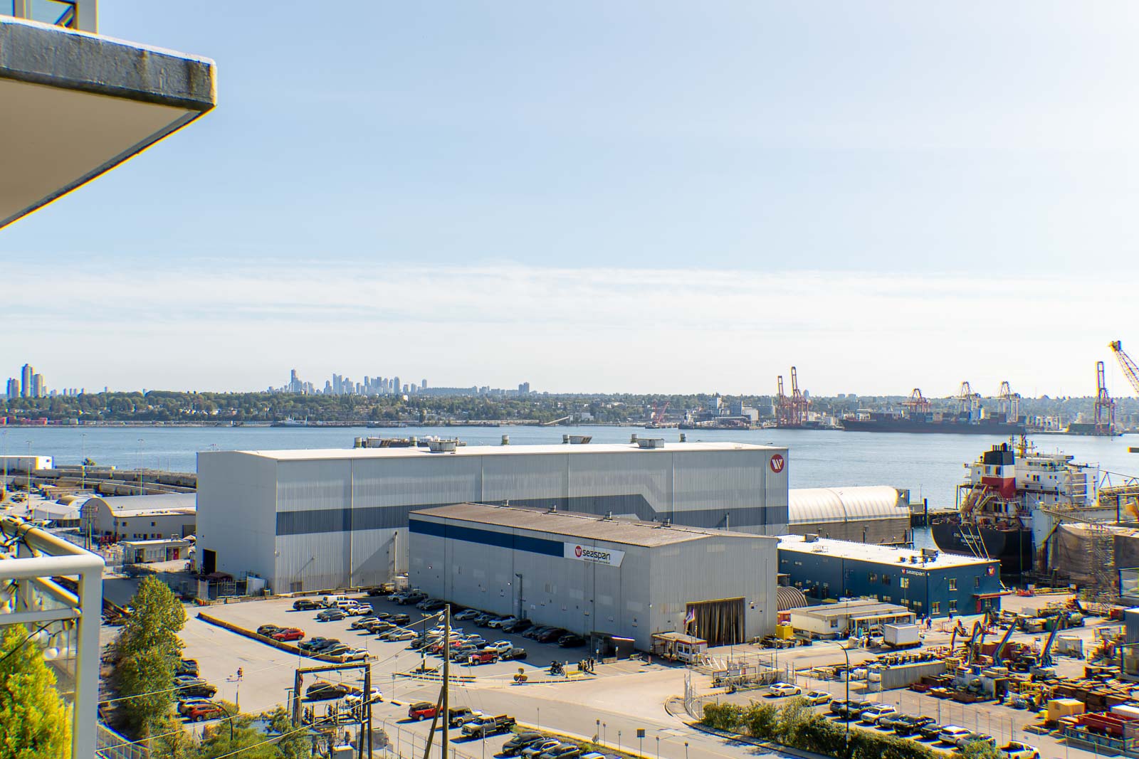 Industrial port with a large warehouse, cars, containers, and ships; Vancouver skyline seen from 188 E Esplanade, clear sky.