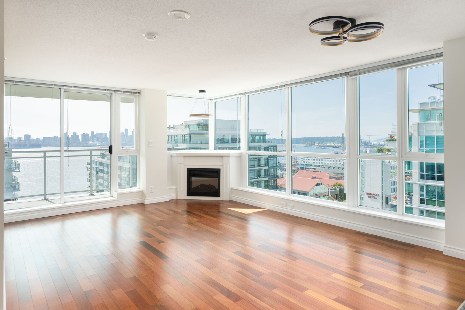 Bright empty living room with hardwood floors, large windows, modern fireplace, and city and water views in North Vancouver.