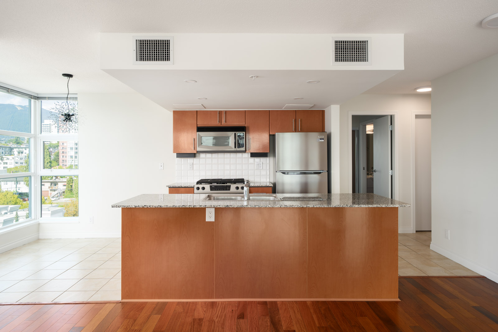 Modern kitchen with wood cabinets, granite island, stainless appliances, and large windows overlooking North Vancouver cityscape.