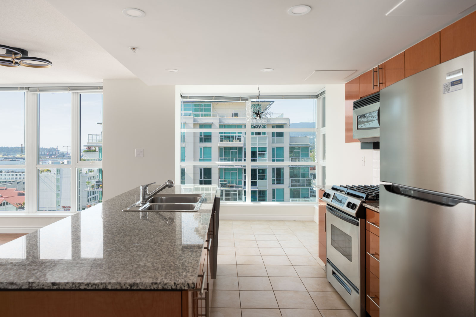 Modern kitchen with granite island, stainless appliances, and large windows overlooking city buildings from 12th floor in North Vancouver.