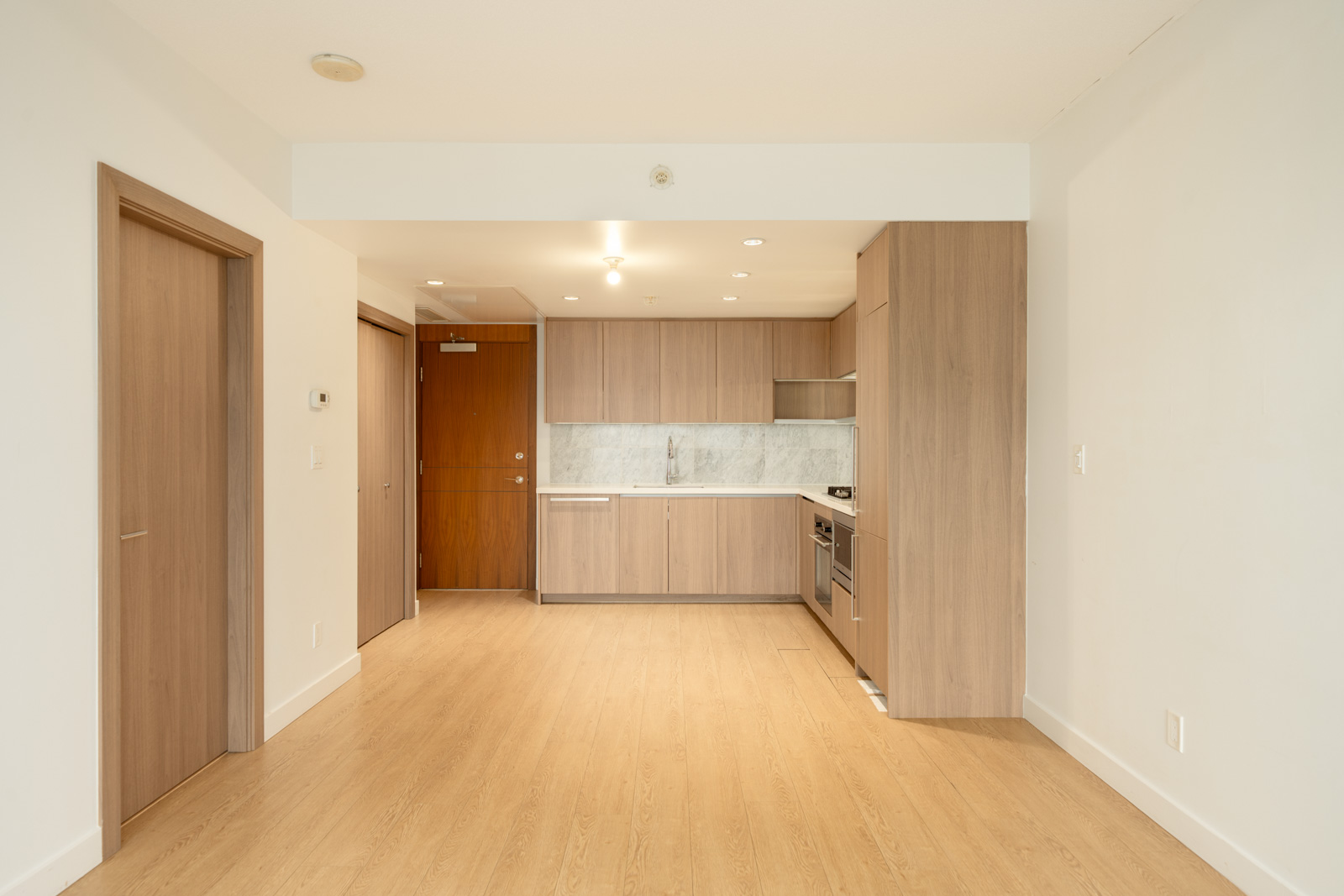 Modern Burnaby kitchen with light wood cabinets, marble backsplash, integrated appliances, wood floors, and two closed doors.
