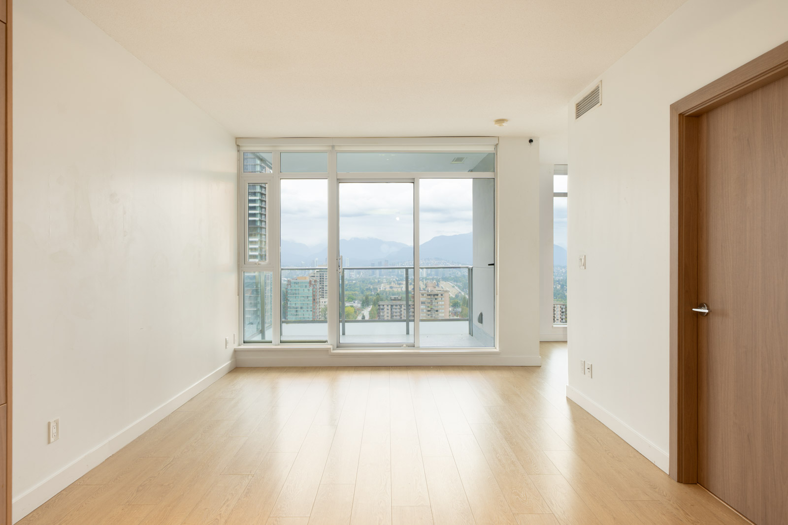 Empty room with light wood floors, white walls, and floor-to-ceiling windows to a balcony overlooking city and mountains in Burnaby.