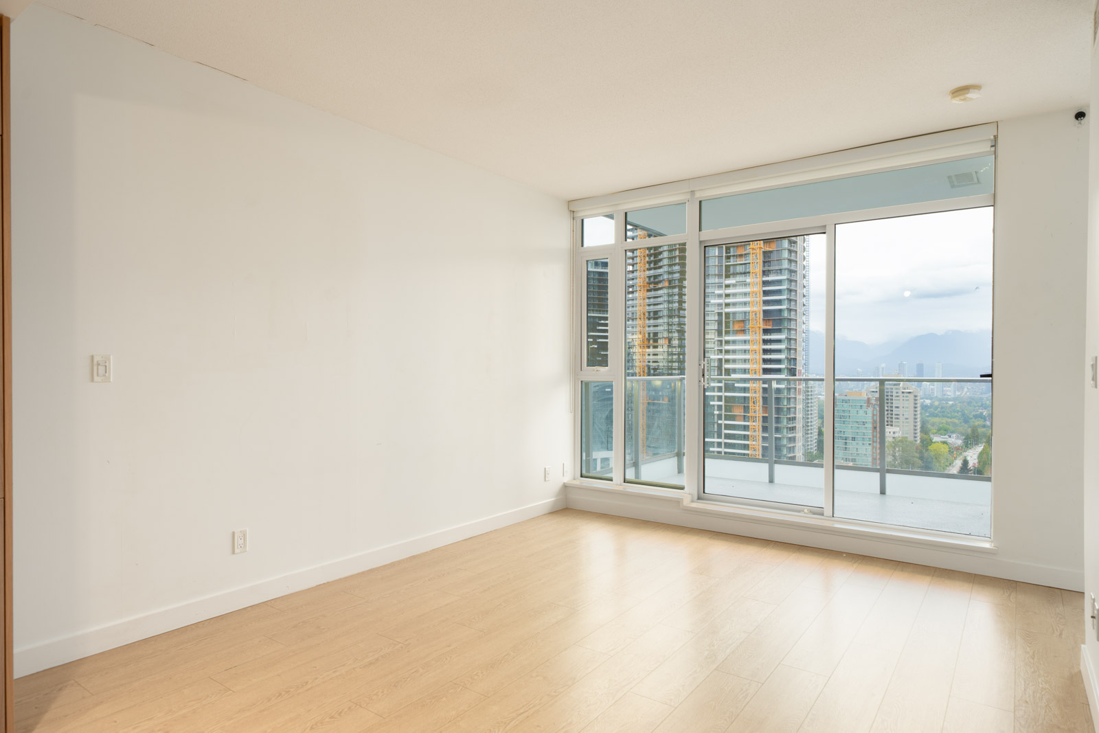 Empty room with light wood floors, white walls, and large windows overlooking city buildings and mountains in Burnaby, on Nelson Ave.