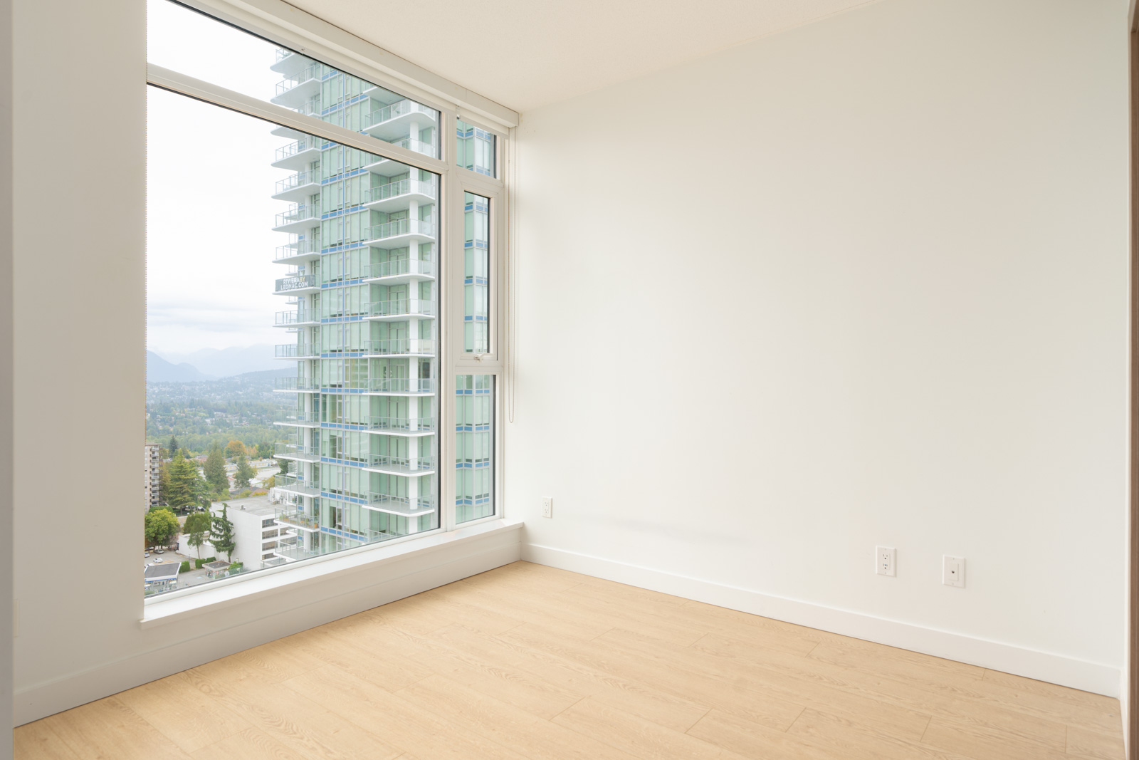 Empty room with light wood floors, white walls, and a large window overlooking city high-rises at 6538 Nelson Ave, Burnaby.