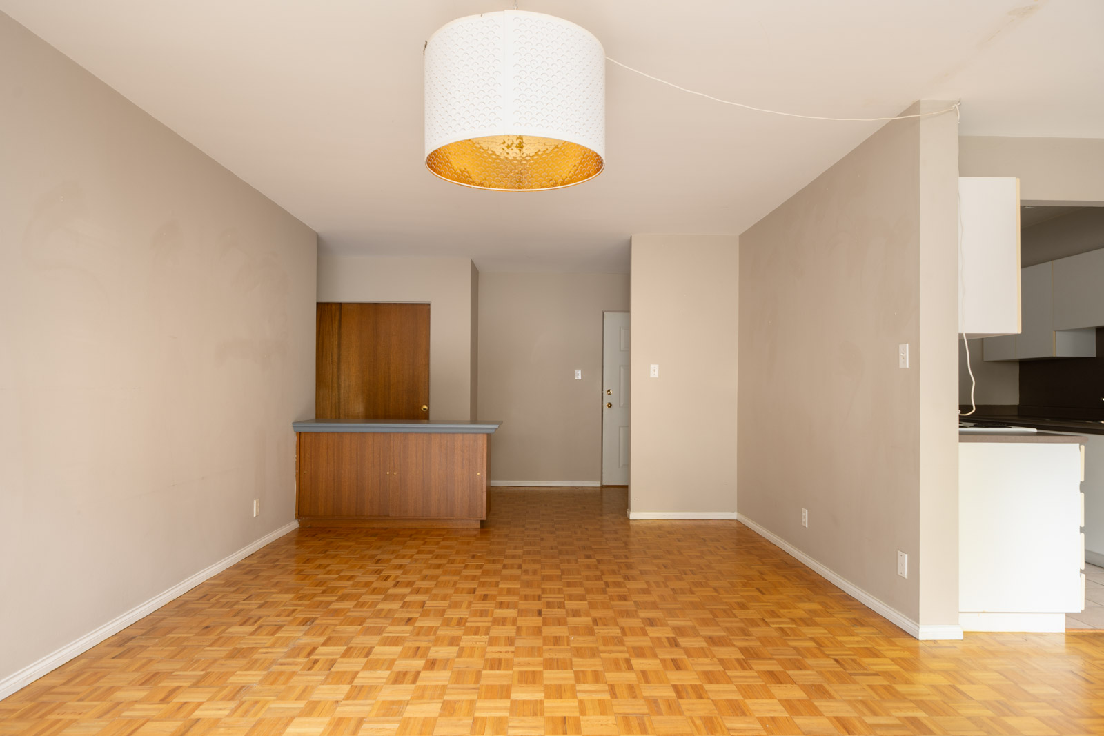 Empty room with parquet floor, beige walls, round ceiling light, wood counter, closed door, and partial kitchen at 1970 W 41st Ave, Vancouver.