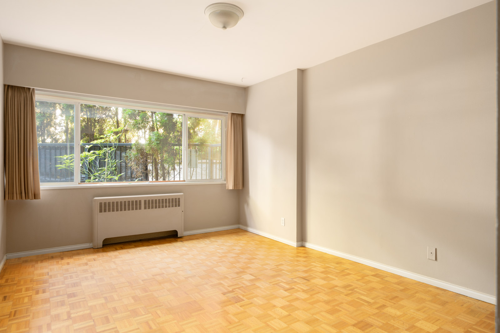 Sunlit empty room at 1970 W 41st Ave, Vancouver with parquet floors, beige walls, large window, and wall heater below.