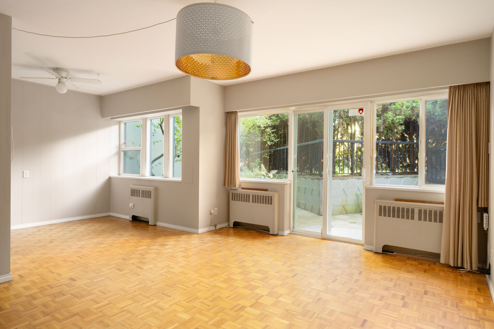 Bright empty room with parquet floors, large windows, beige walls, ceiling fan, and patio access with greenery in Vancouver.